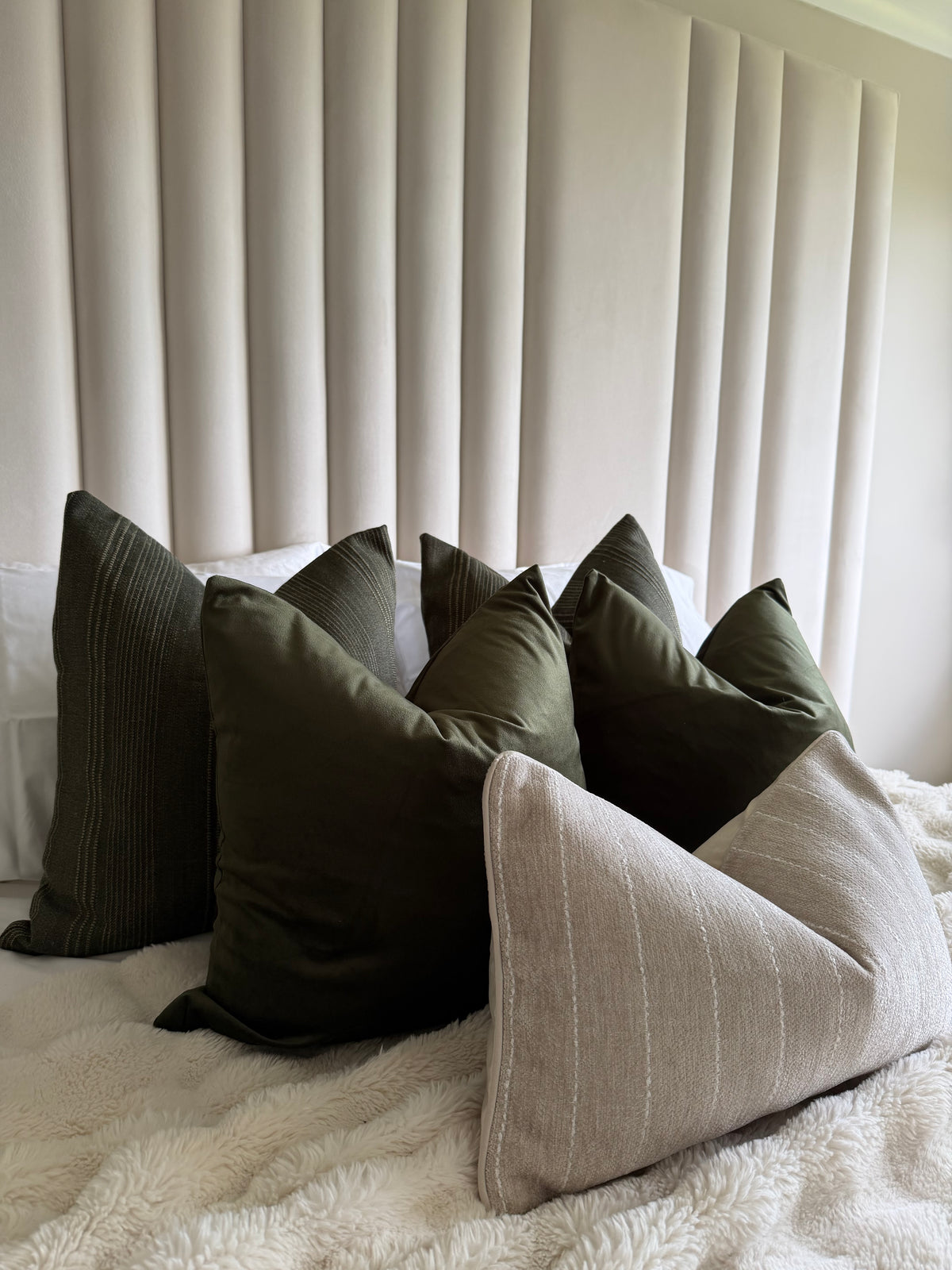 A styled collection of home decor pillows arranged on a bed with a textured cream blanket and a vertical pleated headboard. The pillows include dark olive green, dark gray, and a light beige linen-textured triangle pillow.