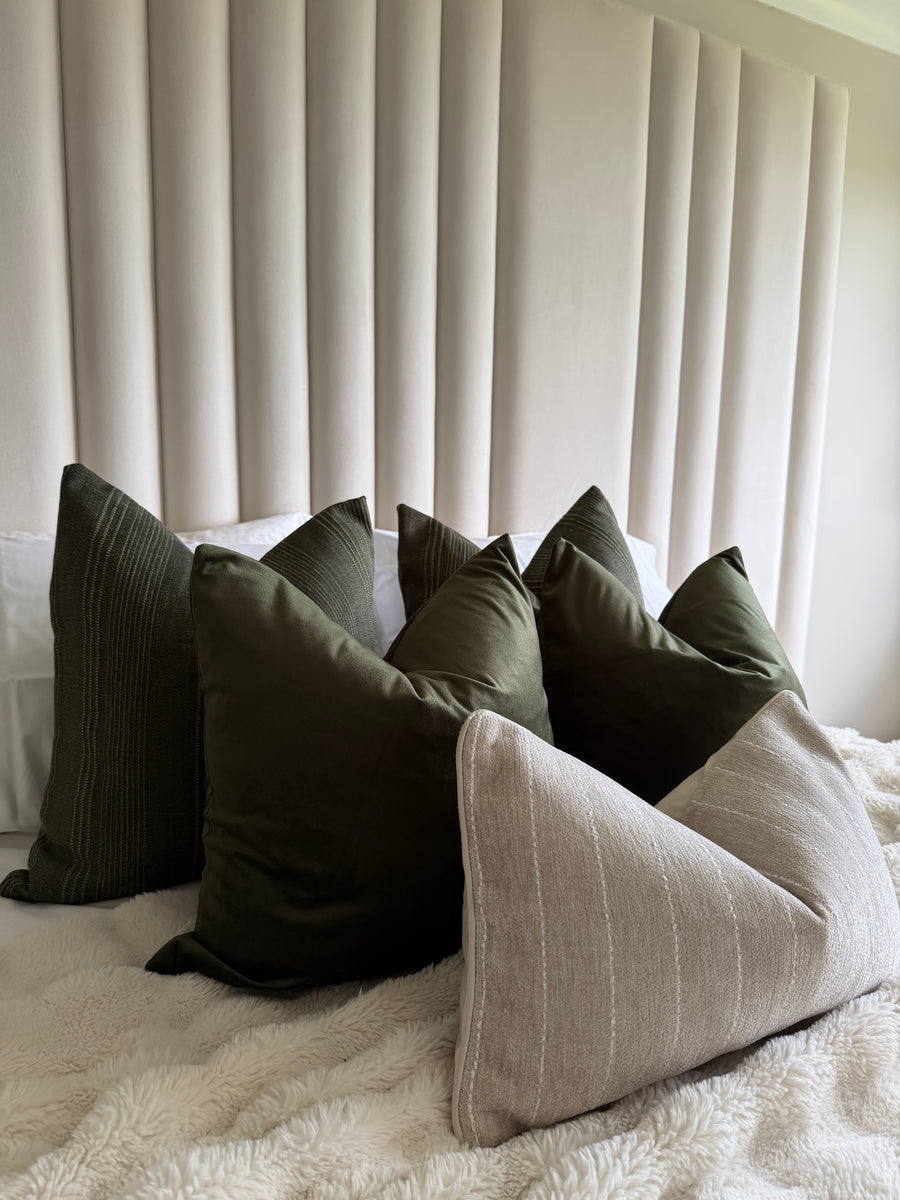 A styled collection of home decor pillows arranged on a bed with a textured cream blanket and a vertical pleated headboard. The pillows include dark olive green, dark gray, and a light beige linen-textured triangle pillow.
