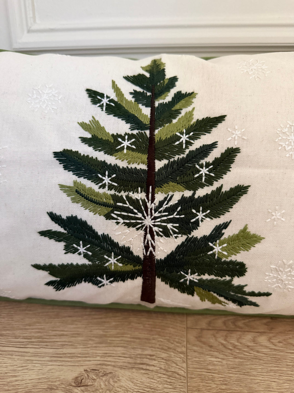 A rectangle white throw pillow featuring an embroidered green Christmas tree with white snowflake details, resting on a light wood floor.
