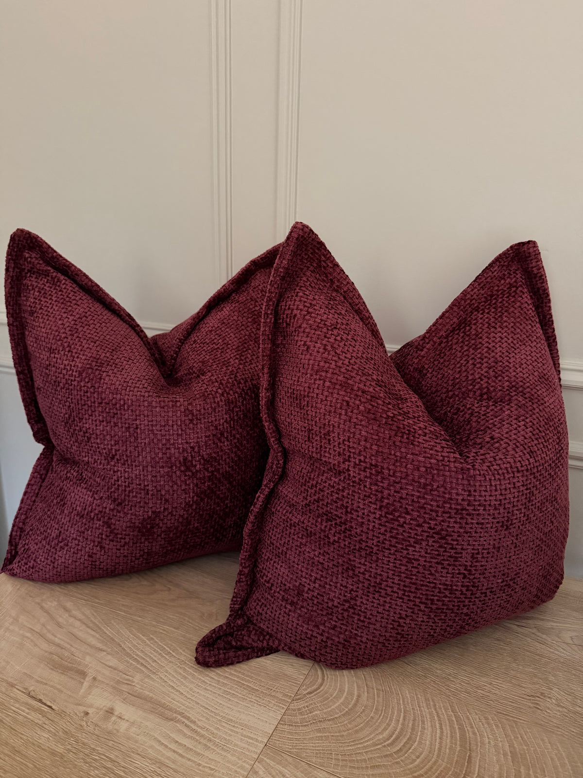 Two textured burgundy throw pillows with a woven, basketweave-like fabric, displayed on a light wood floor against a cream paneled wall.