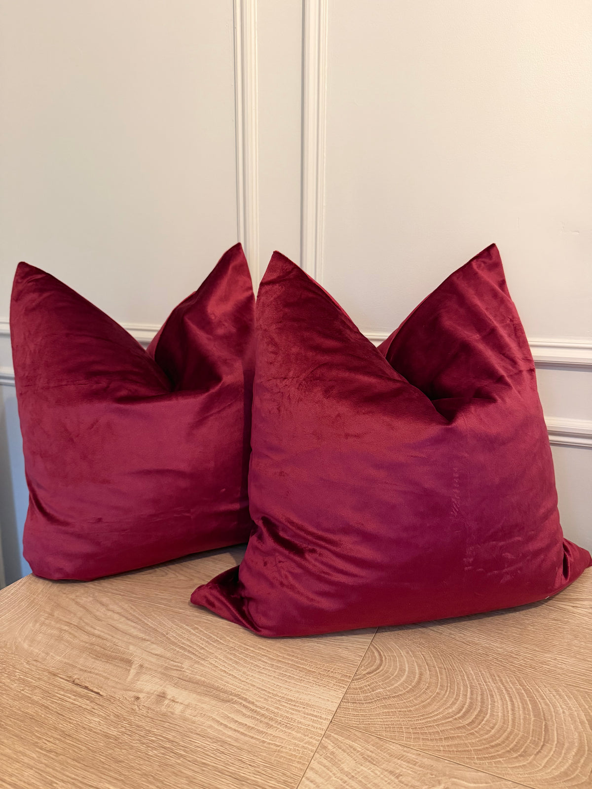 Two plush velvet throw pillows in a deep burgundy red color, styled on a light wood floor against a white paneled wall.